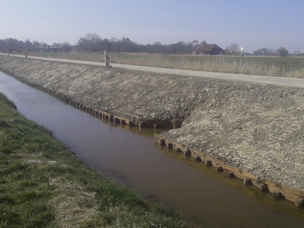 Sanierte Berme mit steilem, mit Holzpfählen gesicherten Uferabschnitt, daneben ein unbefestigter Weg und flaches Ackerland im Hintergrund.
			